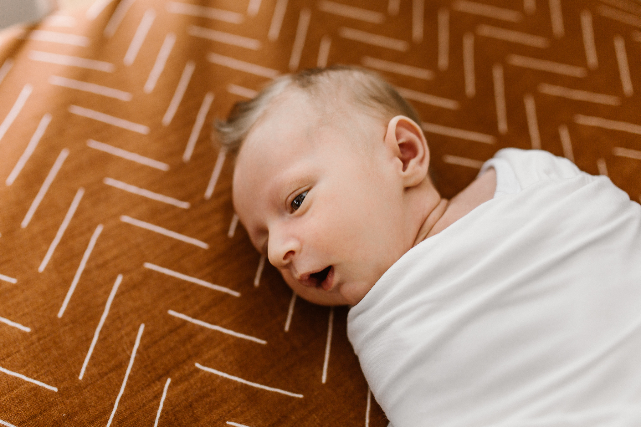 Baby boy swaddled in a white blanket in his crib with window light shining in.